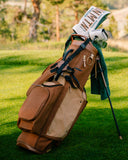 A brown and beige TQR Waxed Canvas golf bag standing on a grassy golf course, with golf clubs and a green towel tucked into the top pocket.