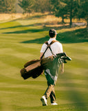 A man on a golf course carrying a brown TQR Waxed Canvas golf bag and clubs on his shoulders.