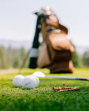 White golf balls with PRACTICE branding on the front, resting on green grass next to wooden tees, with a blurred golf bag and clubs in the background on a golf course.