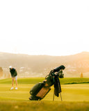A TQR Waxed Canvas golf bag filled with clubs stands on a grassy golf course under warm golden sunlight, with a golfer visible in the background near the horizon.