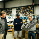 Designer, Seth, holds up a gray WeberState golf bag with white script lettering, while Joel Dahmen, Grant Knudson, and Grant Lordan observe in the Sun Mountain warehouse.