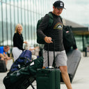 Joel Dahmen pulls a green rolling suitcase while wearing a black long-sleeve shirt with a Sun Mountain logo, gray shorts, a black cap, and a green Sun Mountain backpack. 