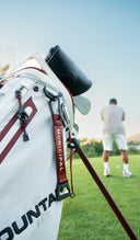 Close-up of a white golf bag with red and black Sun Mountain and MUNICIPAL branding with man teeing off in the background