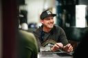 Joel Dahmen wearing a Sun Mountain black baseball cap with white 'SUN.' lettering and a dark long-sleeve shirt with a colorful graphic, seated at a table with cards in a real indoor environment.